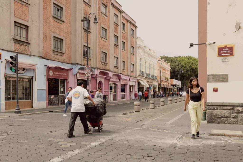 Pedestrians walking on a cobblestone street lined with colorful colonial buildings at the intersection of Avenida 2 Poniente in Puebla, Mexico.